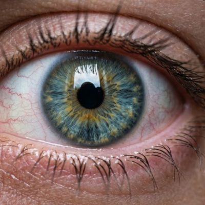Macro photo réaliste d'un œil humain légèrement injecté de sang, vaisseaux fins visibles sur la sclérotique. Regard droit vers l'objectif. Lumière douce, fond noir ou très sombre. Pas de texte. Format 16:9. Rendu clinique mais humain, pas choquant.